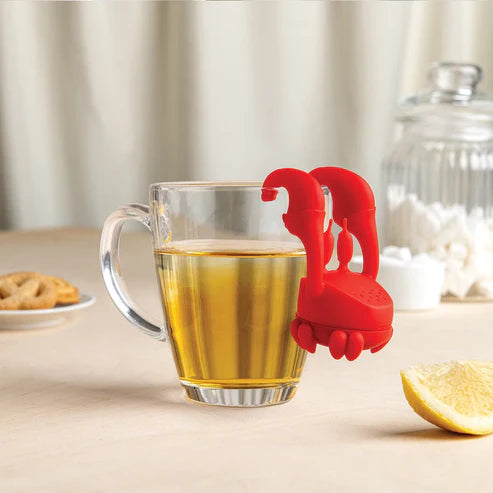 Red tea bag holder in a glass of tea on a table with a lemon and cookies.