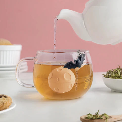 Tea being poured into a glass mug with a silicone tea infuser shaped like a moon with a bat charm.