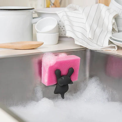 Pink sponge with a black bunny-shaped holder in a sink with soap suds, kitchen setting.
