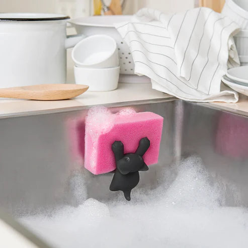 Pink sponge with a black bunny-shaped holder in a sink with soap suds, kitchen setting.